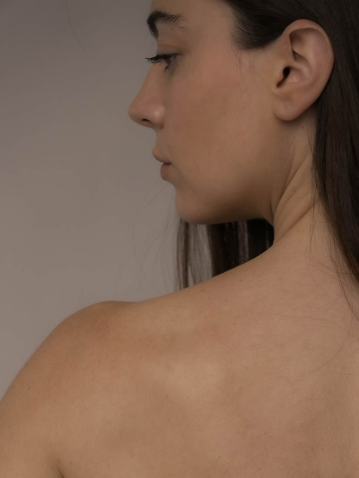 Close-up side profile of a woman's head and bare shoulder against a plain warm gray background.