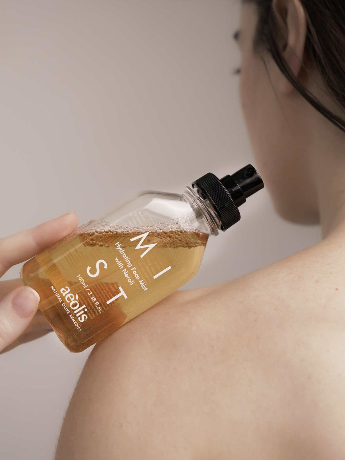 A person holding a clear bottle of Aeolis Face Mist with amber-colored liquid and a black spray cap, positioned near their shoulder against a soft beige background.